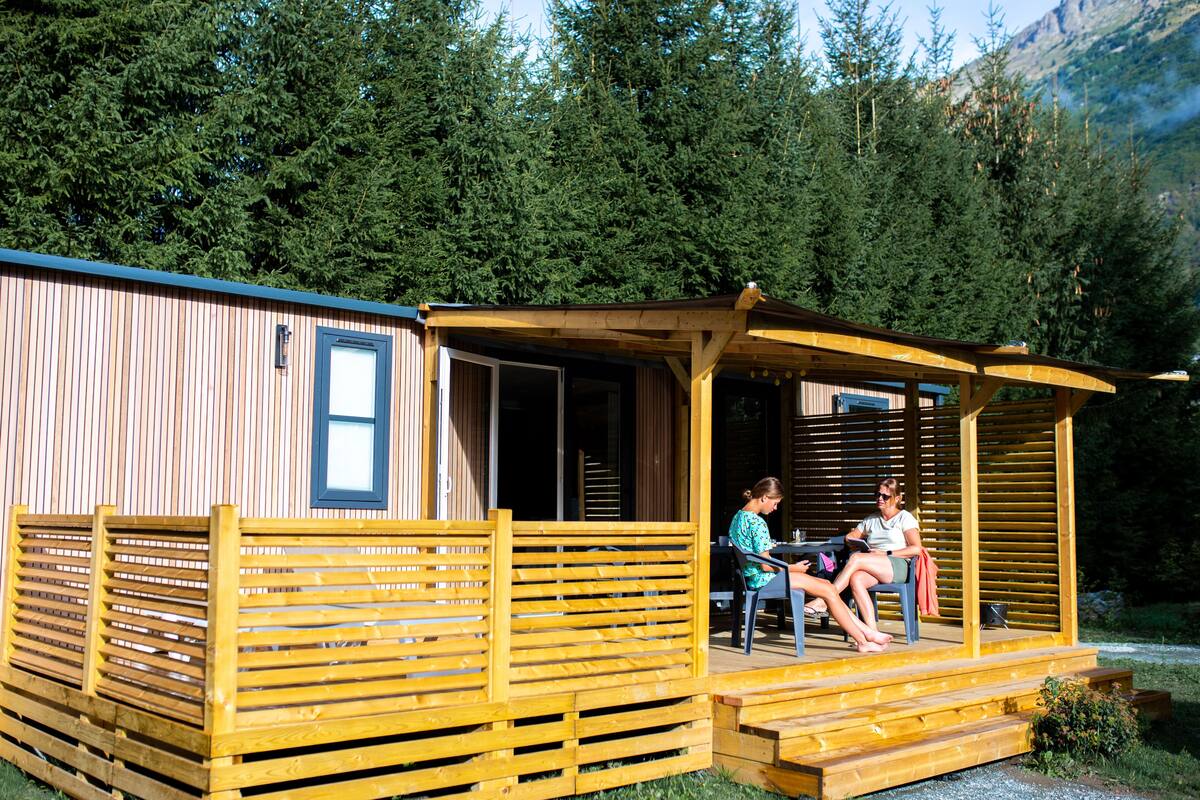 A wooden cabin with a porch, two people seated at a table, and a forested backdrop.