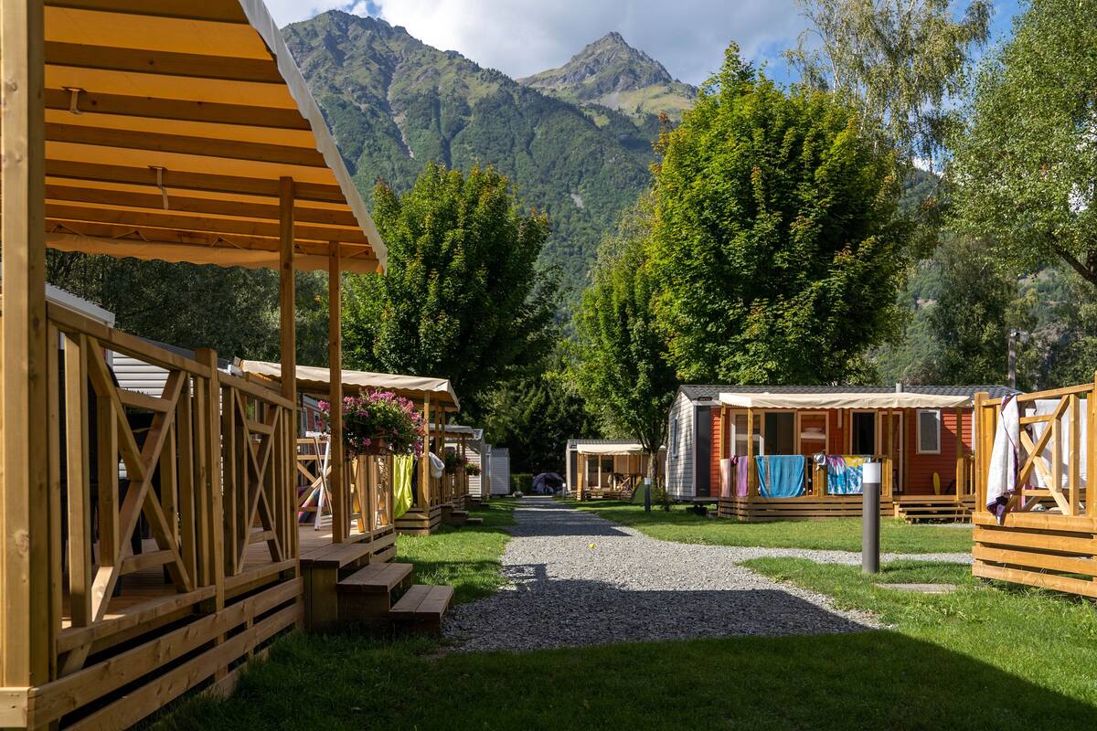 A row of wooden cabins with a gravel path leading through them, set against a backdrop of mountains and trees.