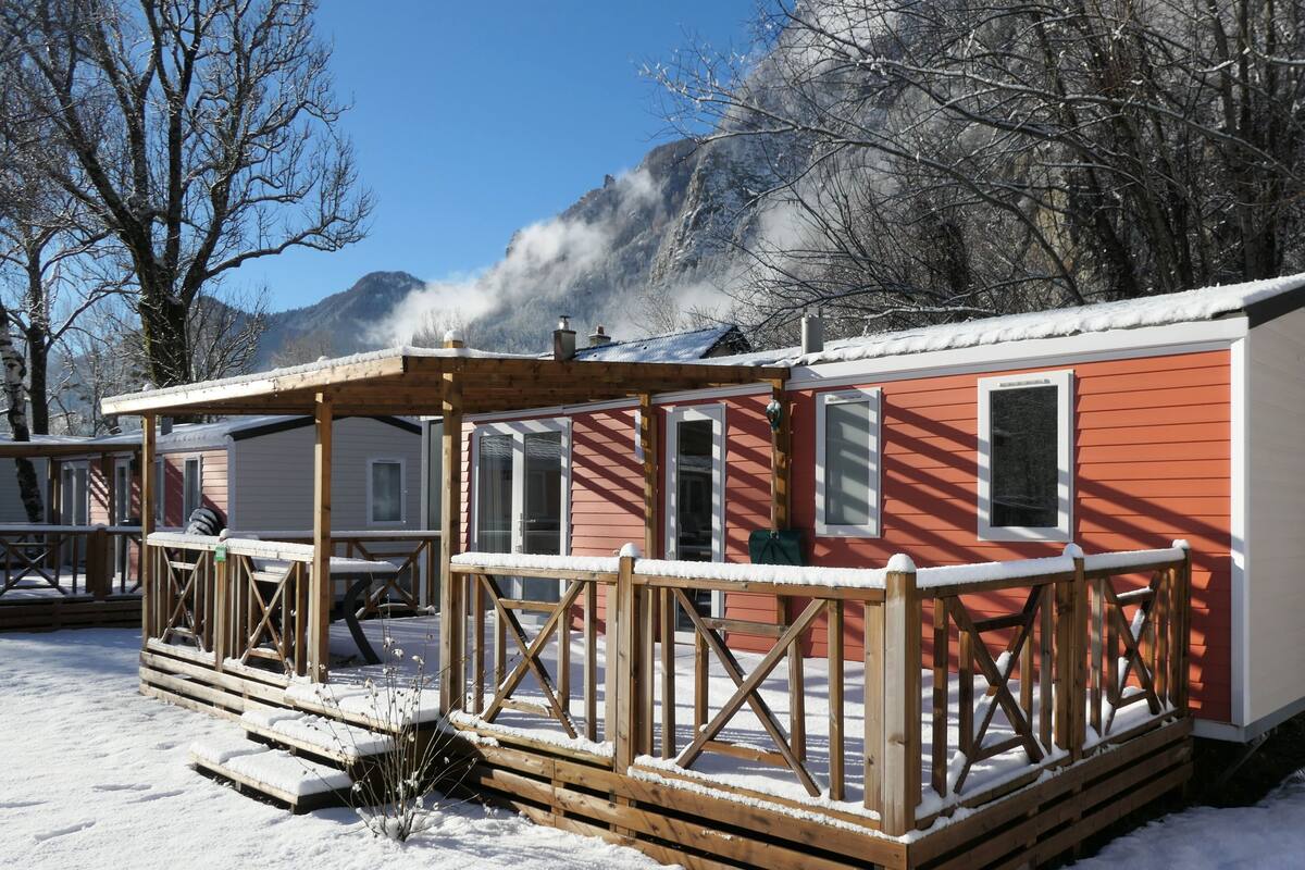A cabin with a wooden deck and a red exterior, surrounded by snow-covered ground and trees.