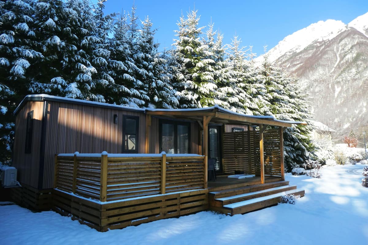 A wooden cabin with a porch and steps, surrounded by snow-covered trees and mountains.