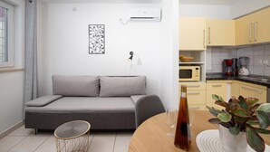 Un espace de vie moderne avec un canapé gris, une petite table ronde, un climatiseur fixé au mur, un four à micro-ondes et une plante en pot.