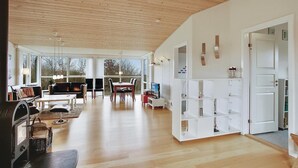 Un vaste espace de vie doté d’un plafond en bois, de grandes fenêtres, d’une table à manger et d’une étagère blanche.