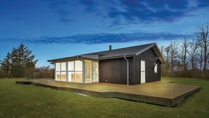 Une maison moderne dotée d’une grande baie vitrée, d’une terrasse en bois et d’une façade de couleur sombre.