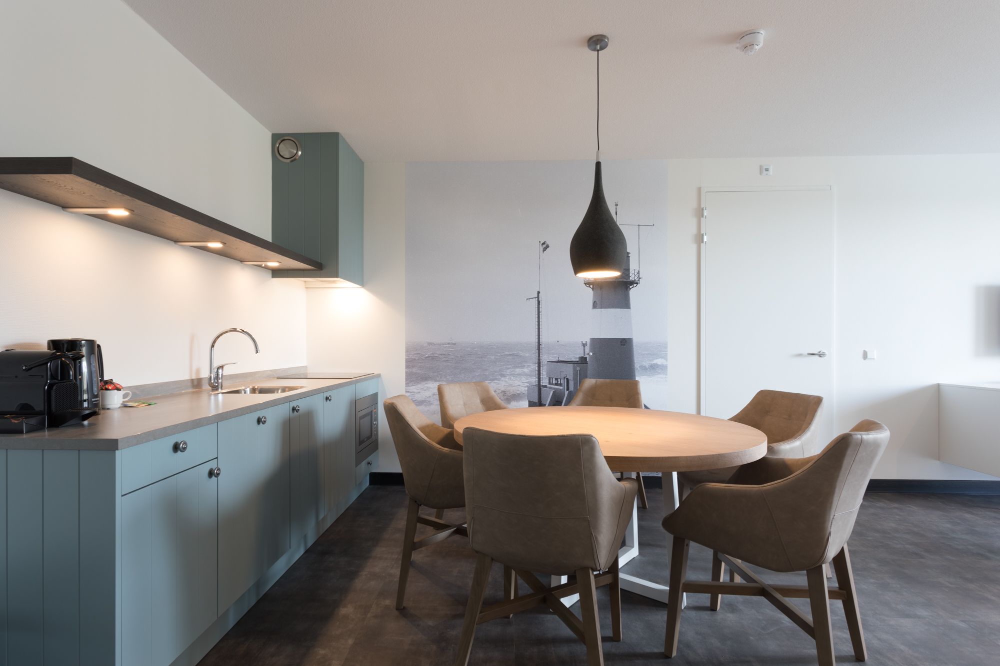 A modern kitchen with a dining area, featuring a large island, pendant lighting, and a view of the sea.
