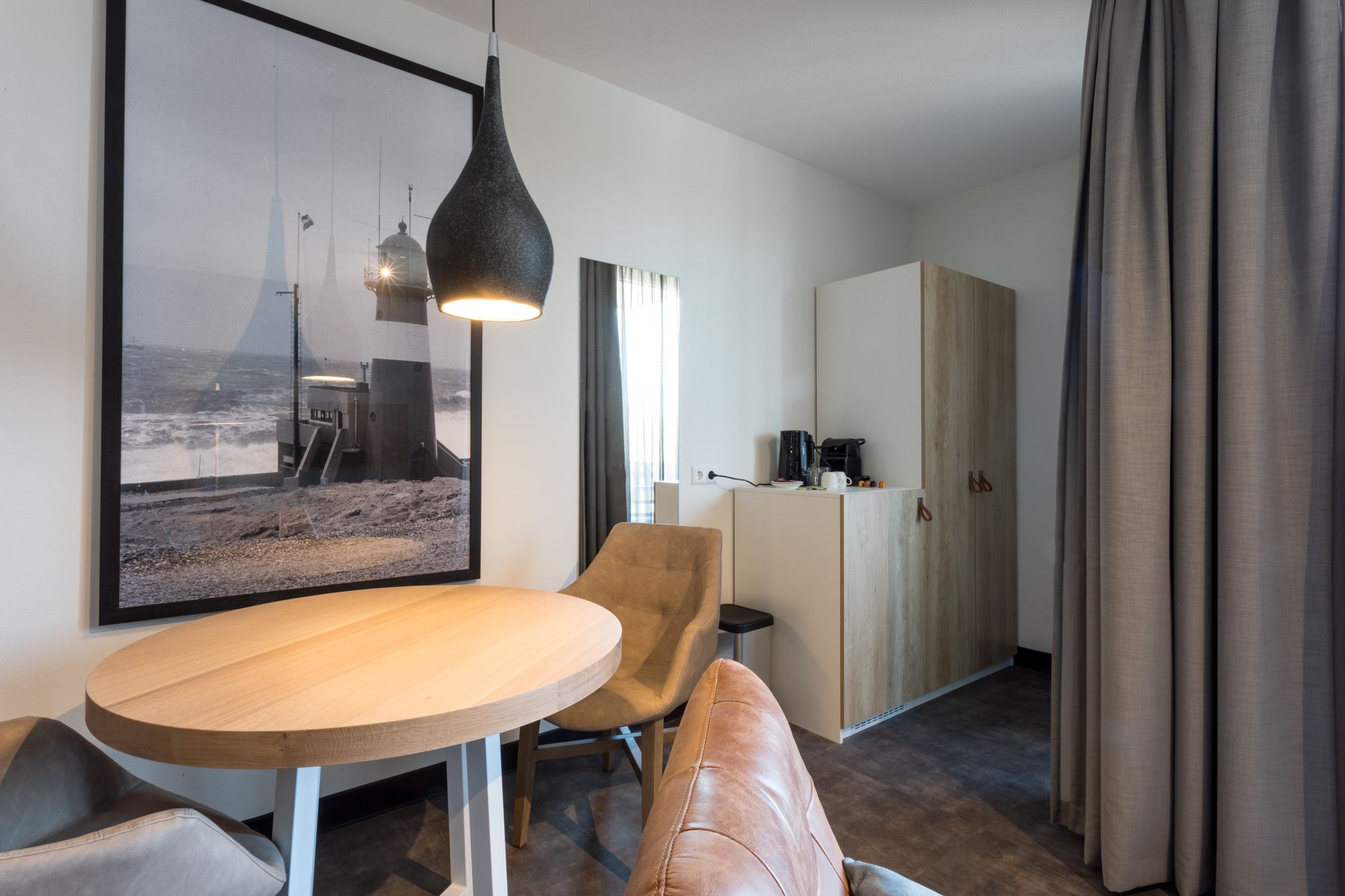A modern hotel room with a round wooden table, a leather sofa, a framed picture of a coastal scene, a pendant light, and a kitchenette.