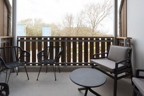 Een balkon met een houten hekwerk, twee rieten stoelen, een comfortabele stoel en een klein rond tafeltje.