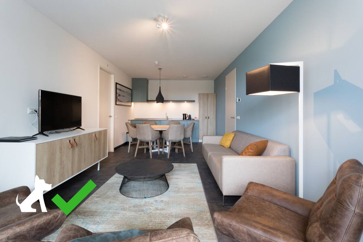 Un salon moderne avec un canapé, une table basse et des fauteuils en cuir. On aperçoit un téléviseur posé sur un meuble, un coin repas avec des chaises, et une cuisine à l’arrière-plan.