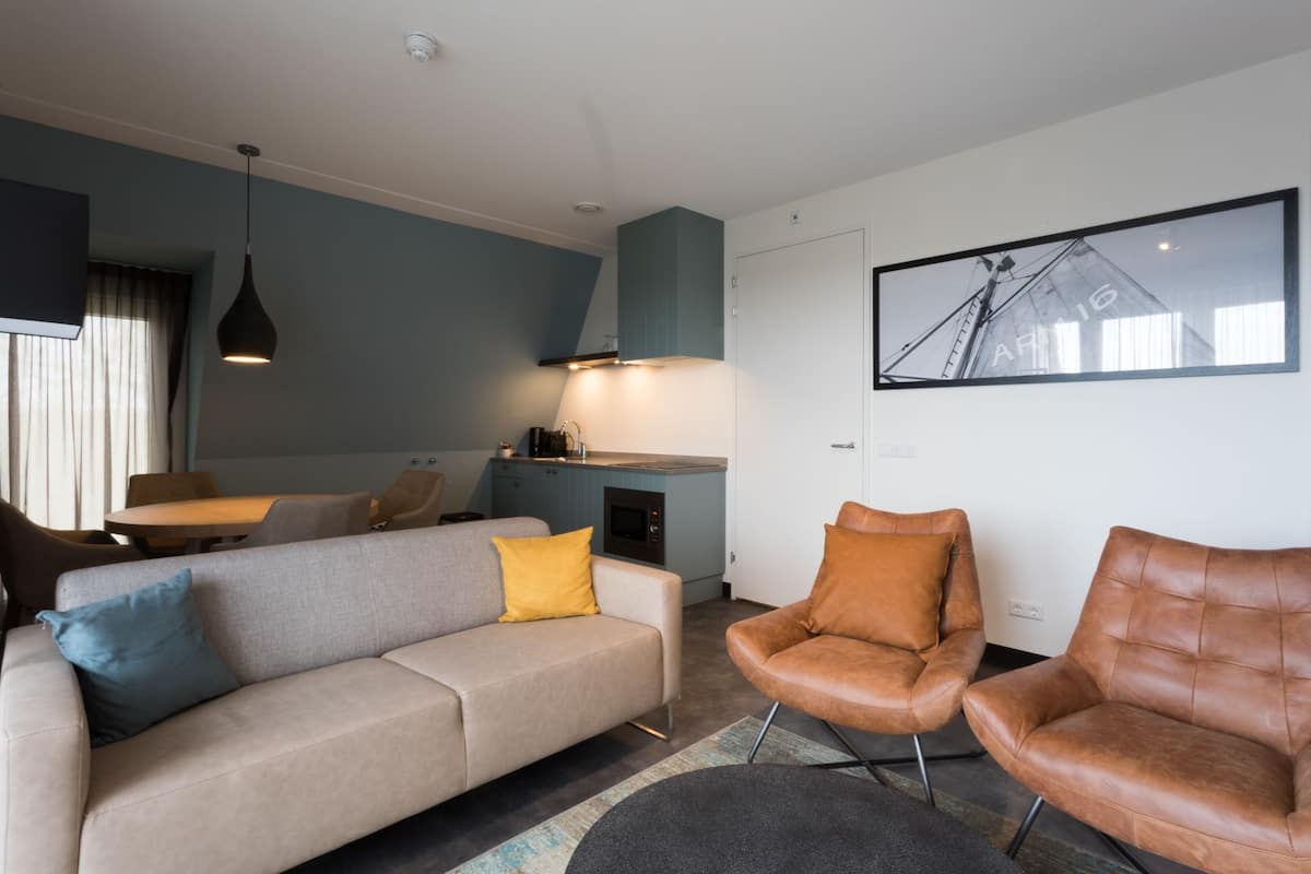 Un salon moderne comprenant un canapé, deux fauteuils en cuir, un coin repas avec une table et des chaises, un coin cuisine et un tableau encadré au mur.
