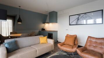 Ein modernes Wohnzimmer mit einer Couch, zwei Ledersesseln, einer Essecke mit Tisch und Stühlen, einer Küchenzeile und einem gerahmten Bild an der Wand.