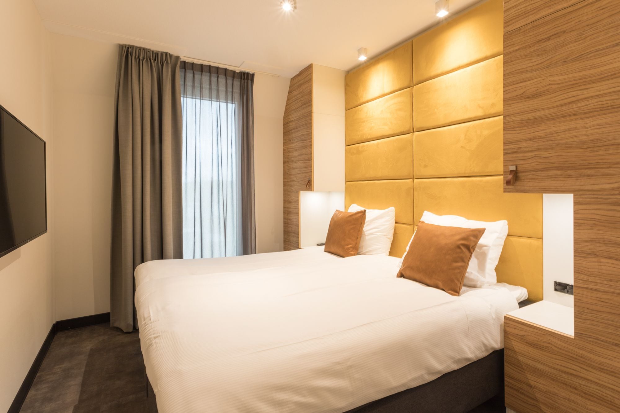 A modern hotel room with a large bed, wooden headboard, and a flat-screen TV.