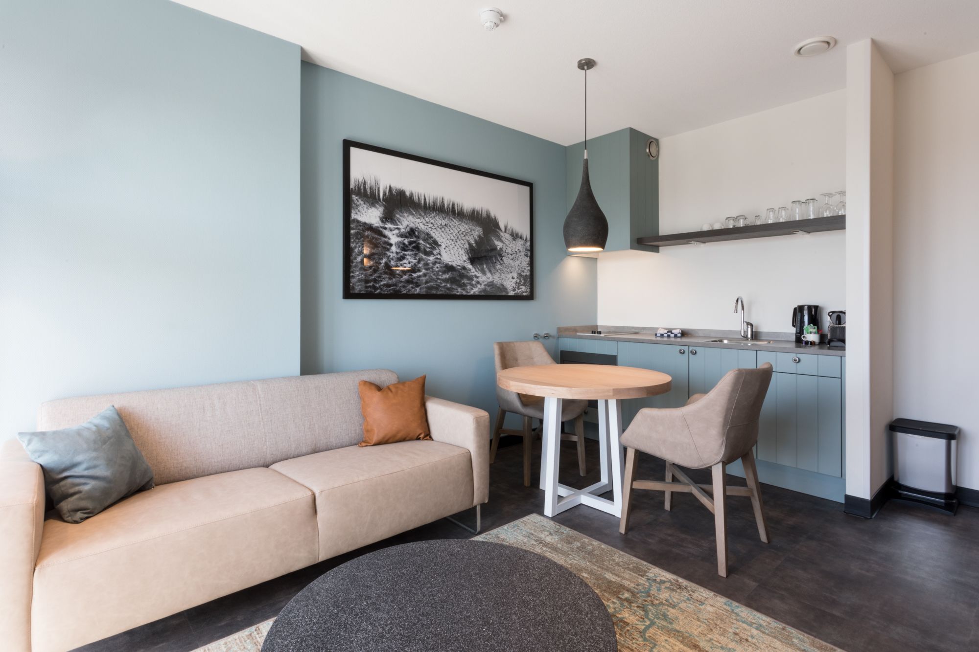 Un salon moderne avec un canapé beige, une table ronde et deux chaises. On aperçoit également un coin cuisine équipé d’un évier, de placards et d’une machine à café.