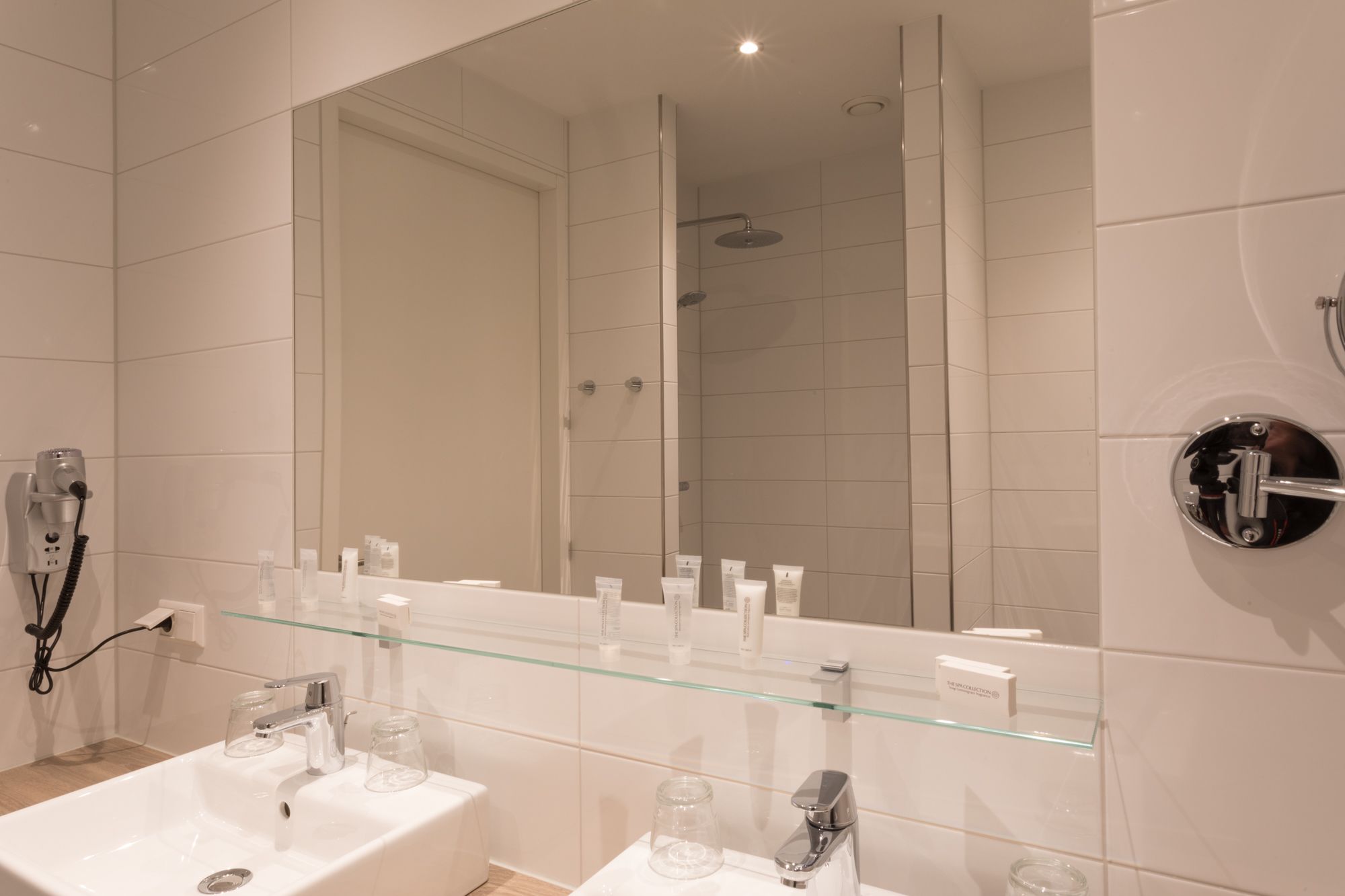 Een moderne badkamer met twee wastafels, een glazen plank met toiletartikelen, een grote spiegel en een douchehoek.