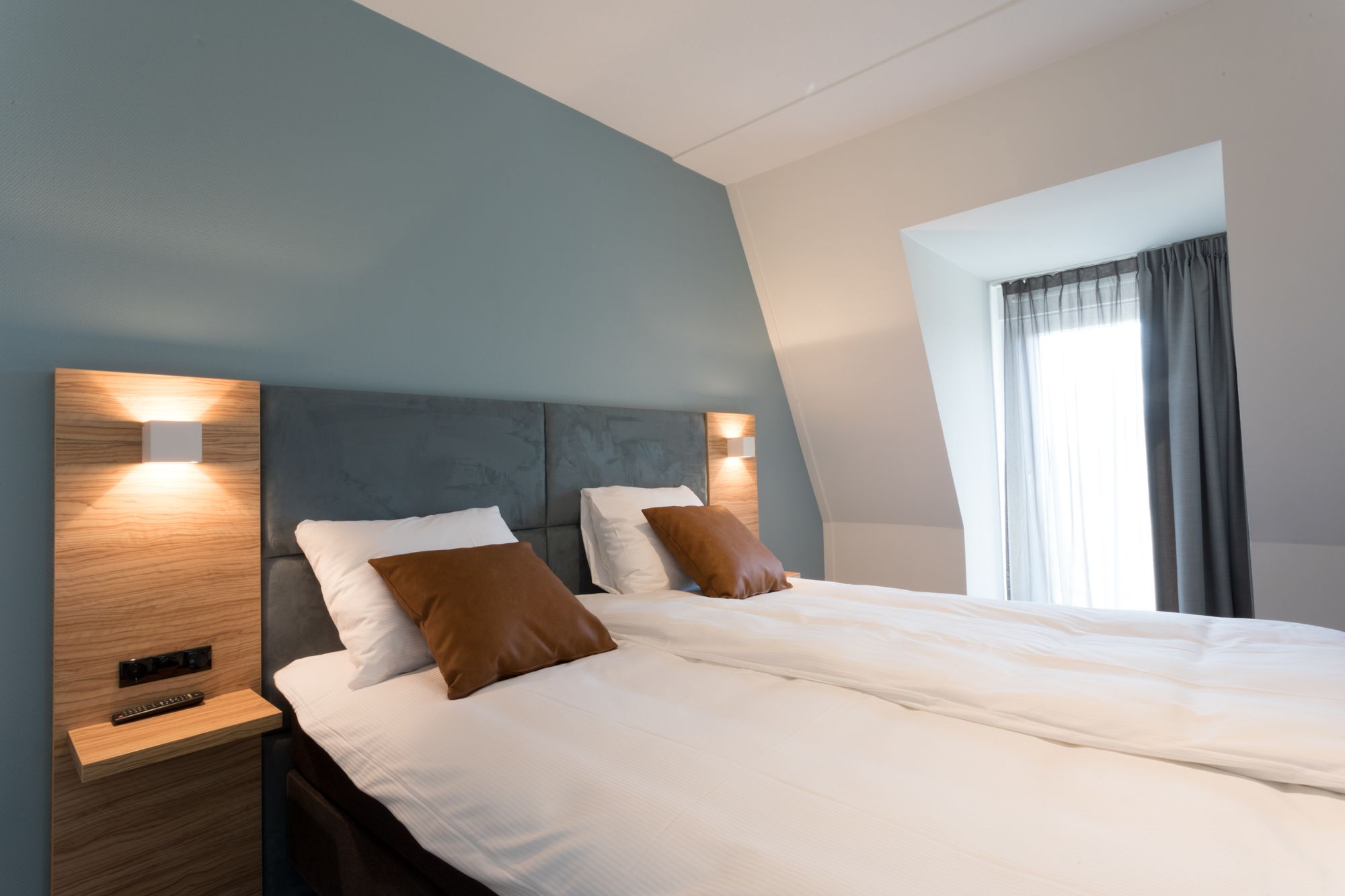 Una habitación de hotel moderna con una cama grande, dos mesitas de noche y un ventanal con cortinas.
