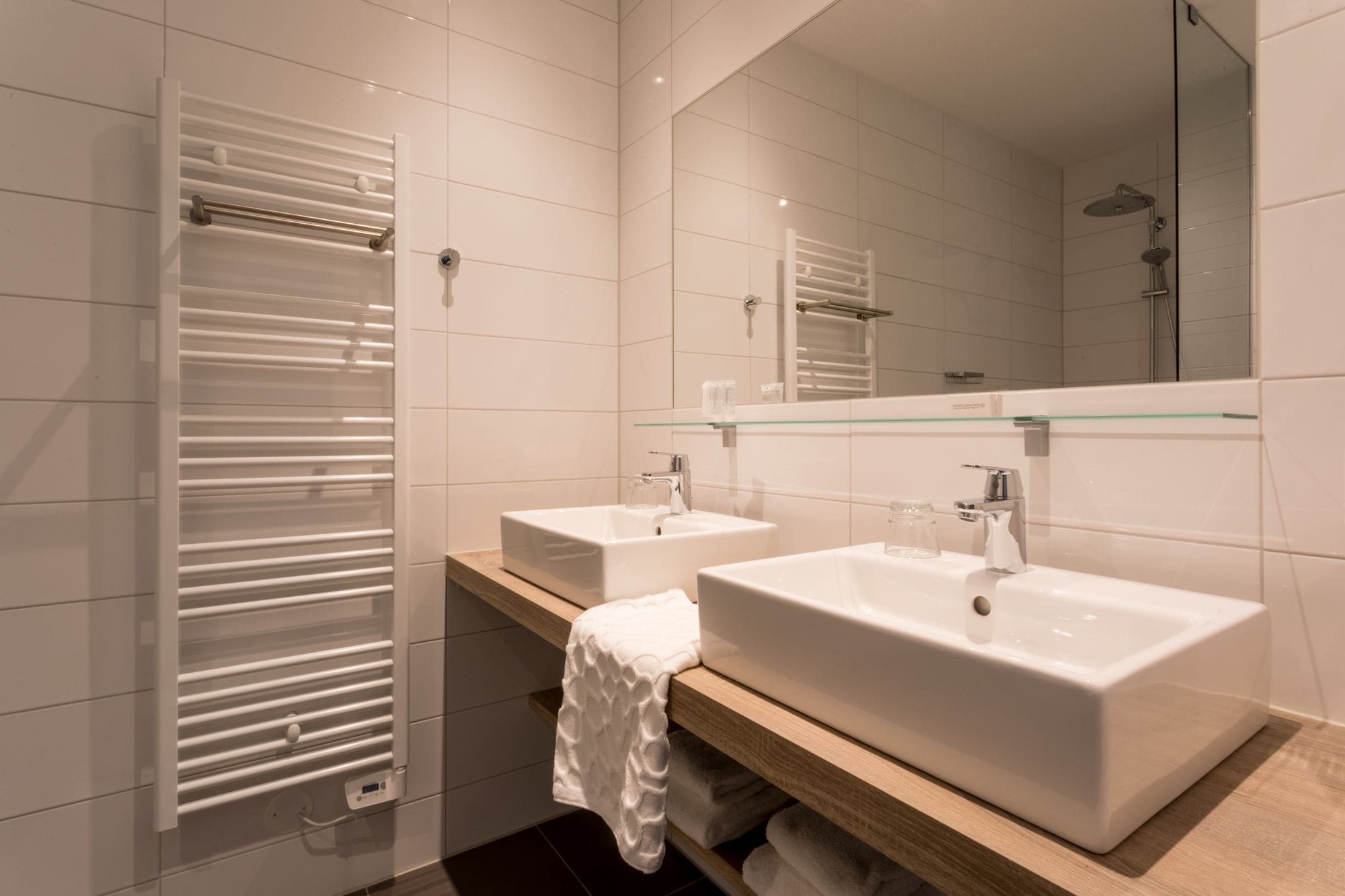 A modern bathroom with two sinks, a glass shelf, and a towel rack.