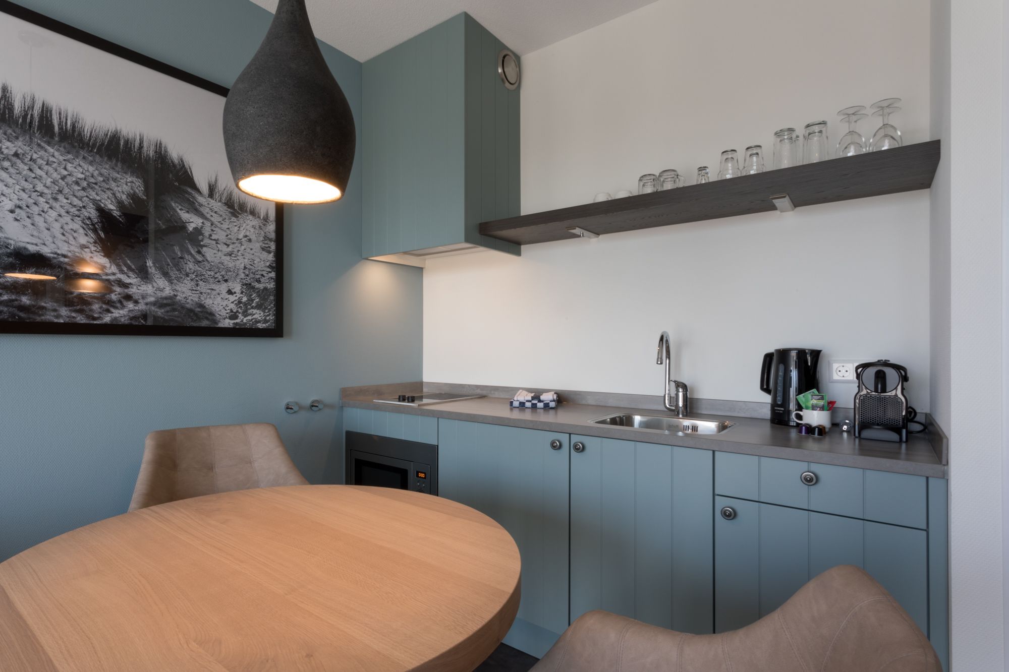 A modern kitchen with a round wooden table, two chairs, a built-in oven, a sink, and a coffee maker.