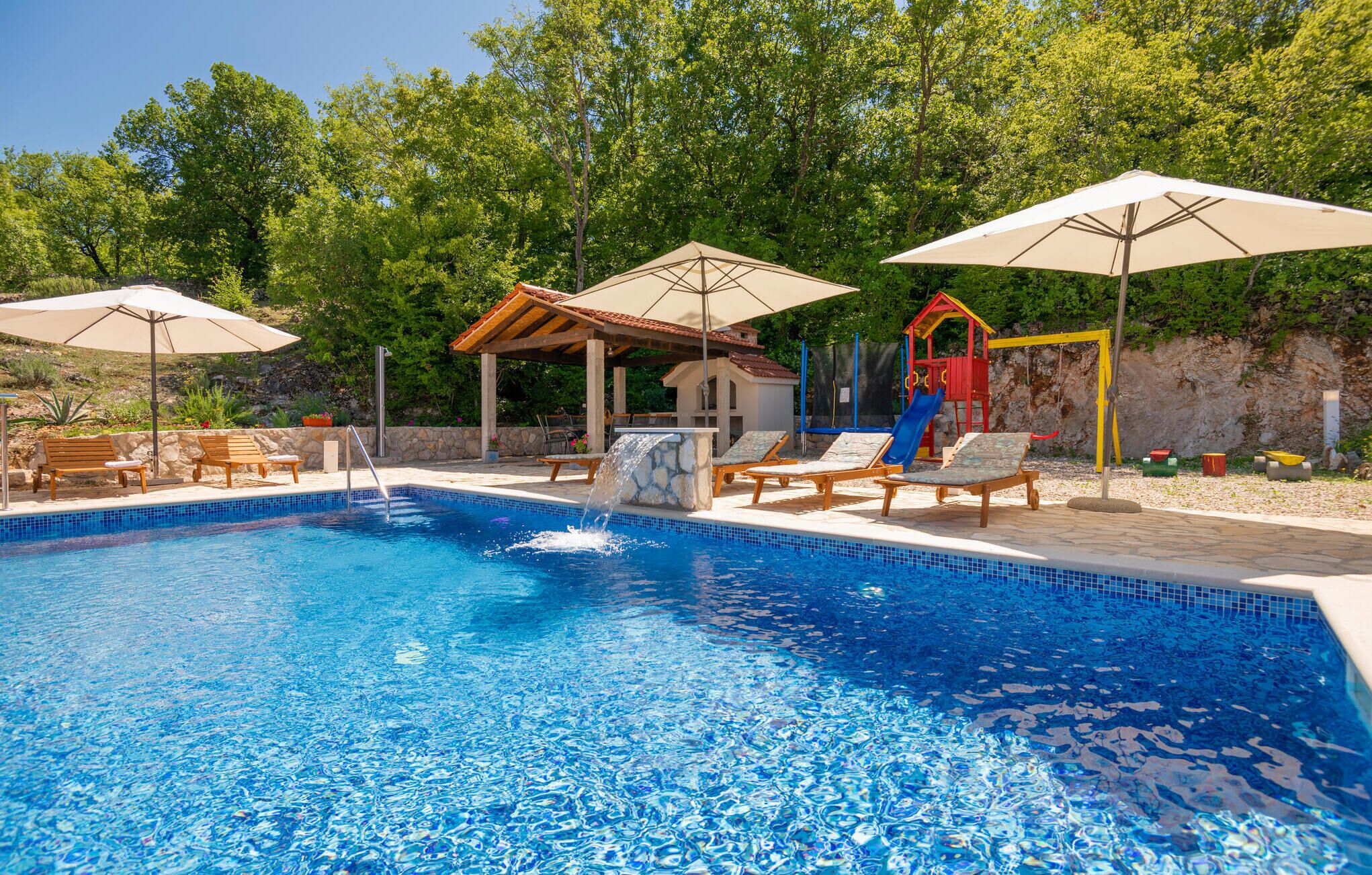 A swimming pool with lounge chairs and umbrellas, a children's play area, and a stone wall.