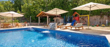 A swimming pool with lounge chairs and umbrellas, a children's play area, and a stone wall.