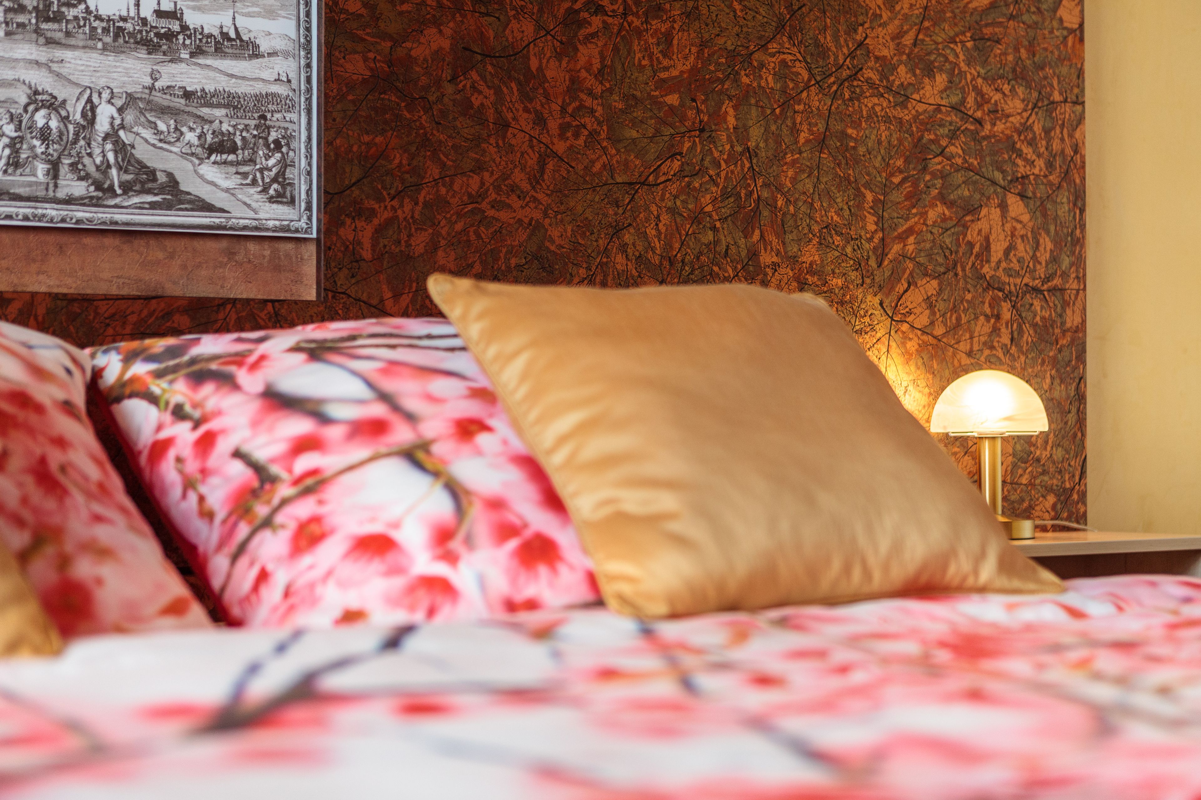 Un lit avec une literie à motifs floraux et un oreiller doré, un tableau accroché au mur et une table de chevet avec une lampe.