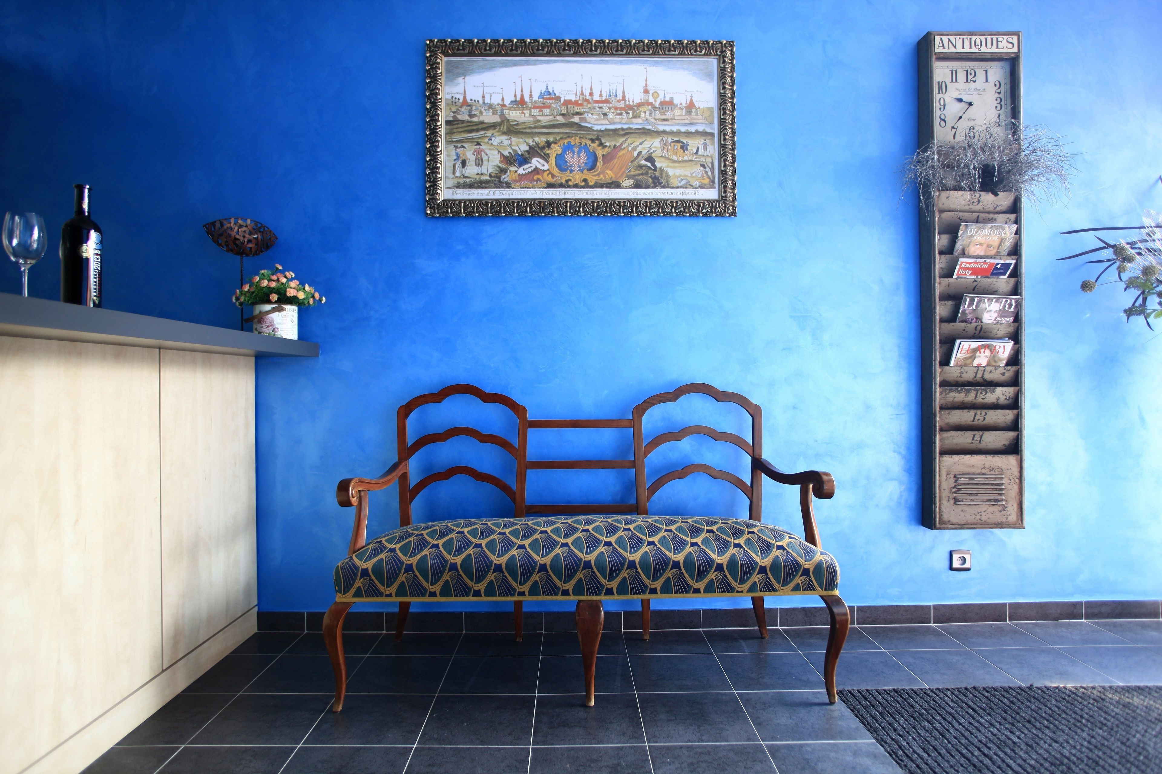 A blue wall with a framed painting, a wooden bench, a shelf with decorative items, and a vertical display of postcards.