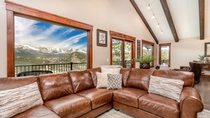 A spacious living room with a large brown leather sectional sofa, a view of snow-capped mountains, and a wooden ceiling with exposed beams. - Breiding Upper Luxury Vacation At Windcliff 2 Bedroom Home by RedAwning (Estes Park)