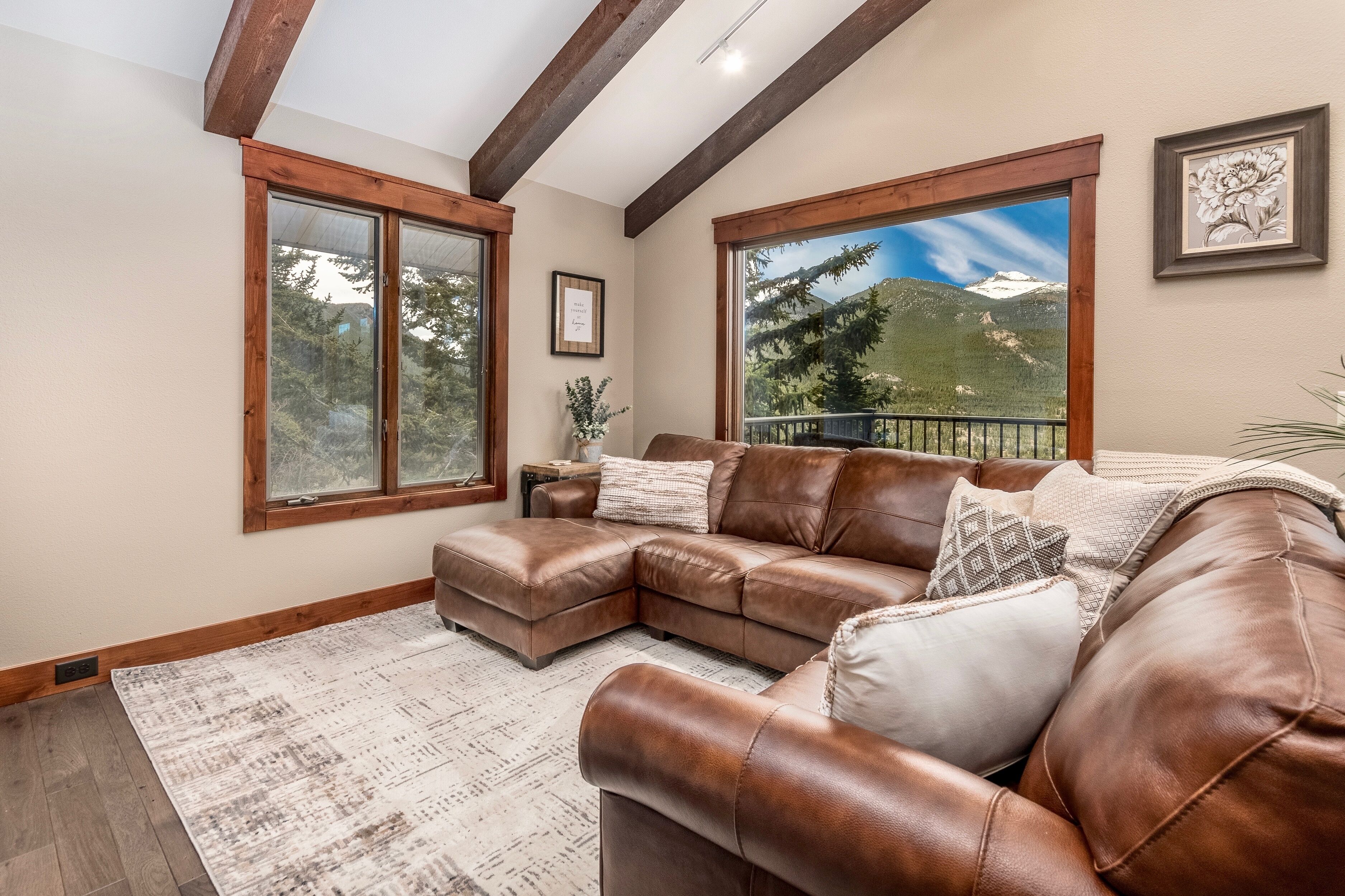 A spacious living room with a brown leather sectional sofa, large windows offering mountain views, and a wooden ceiling.