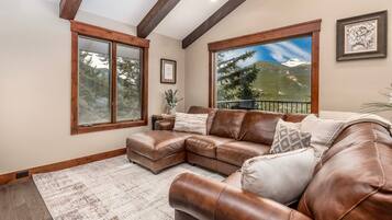 A spacious living room with a brown leather sectional sofa, large windows offering mountain views, and a wooden ceiling.