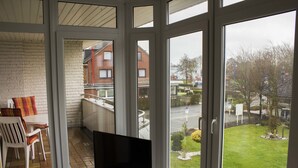 A balcony with a TV, a table, and chairs, overlooking a residential area with houses and a lawn. - Hotel Am Friesenstrand (Butjadingen)