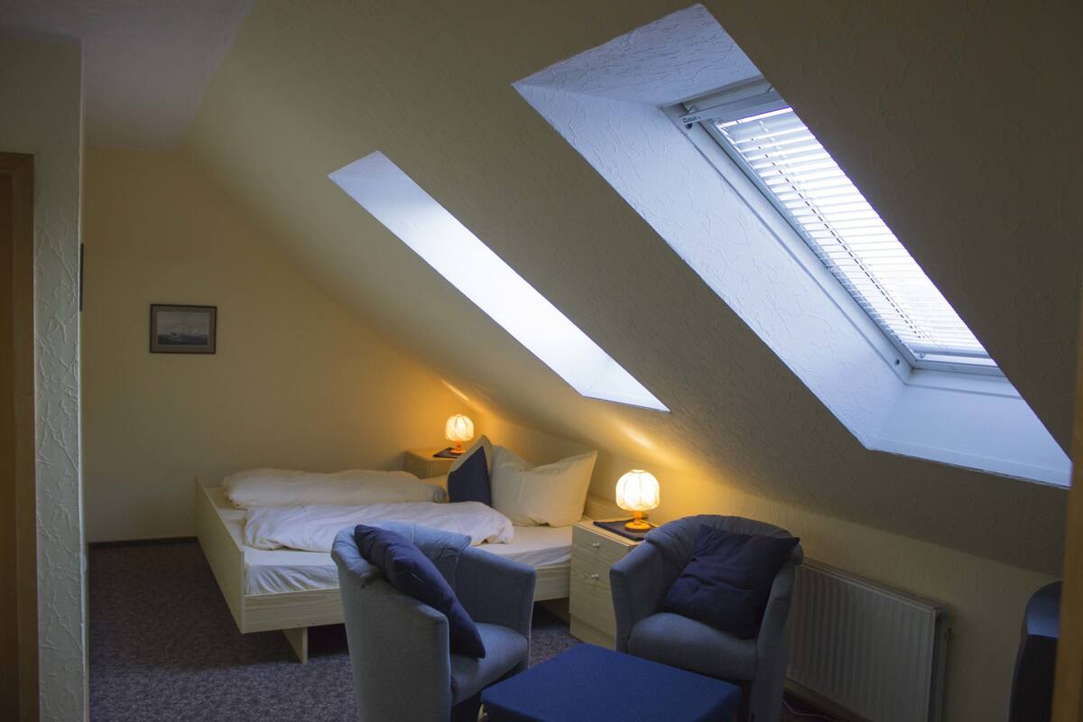 A small, cozy room with a sloped ceiling, a bed, a desk, a chair, and a skylight.