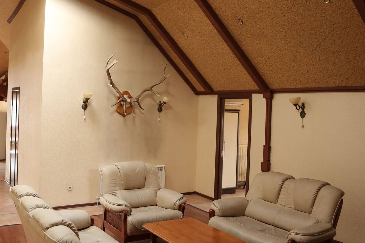 A spacious loft-style room with a high ceiling, wooden beams, and a large window. There is a sofa set and a wooden coffee table.