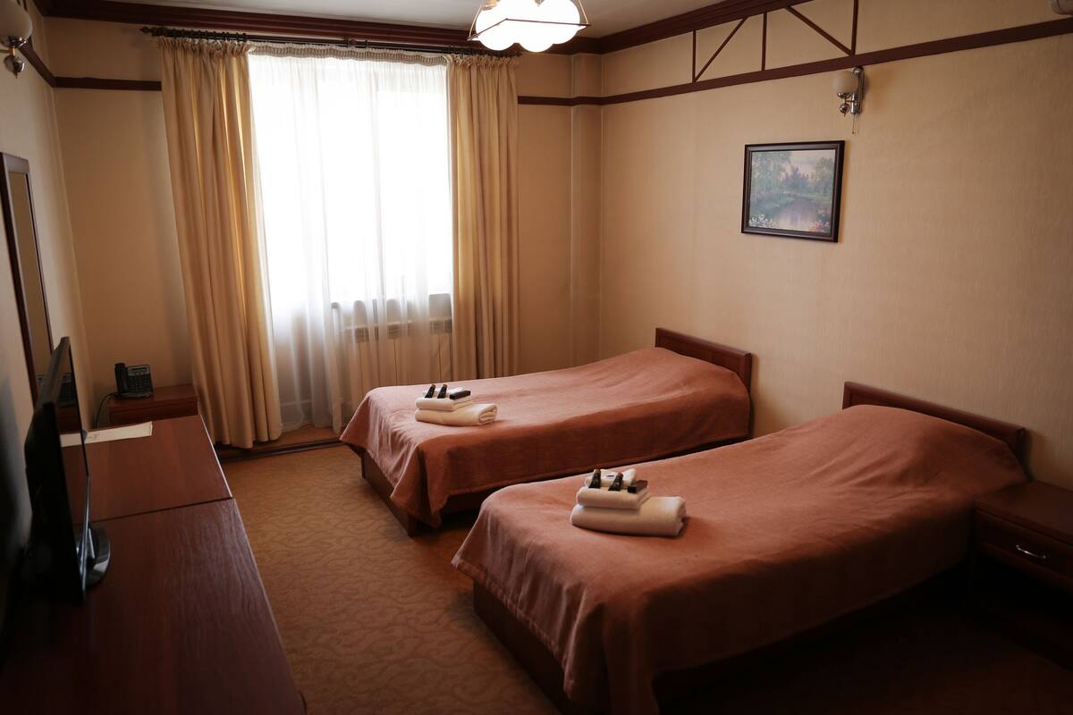 A hotel room with two single beds, a desk, a chair, a telephone, and a window with curtains.