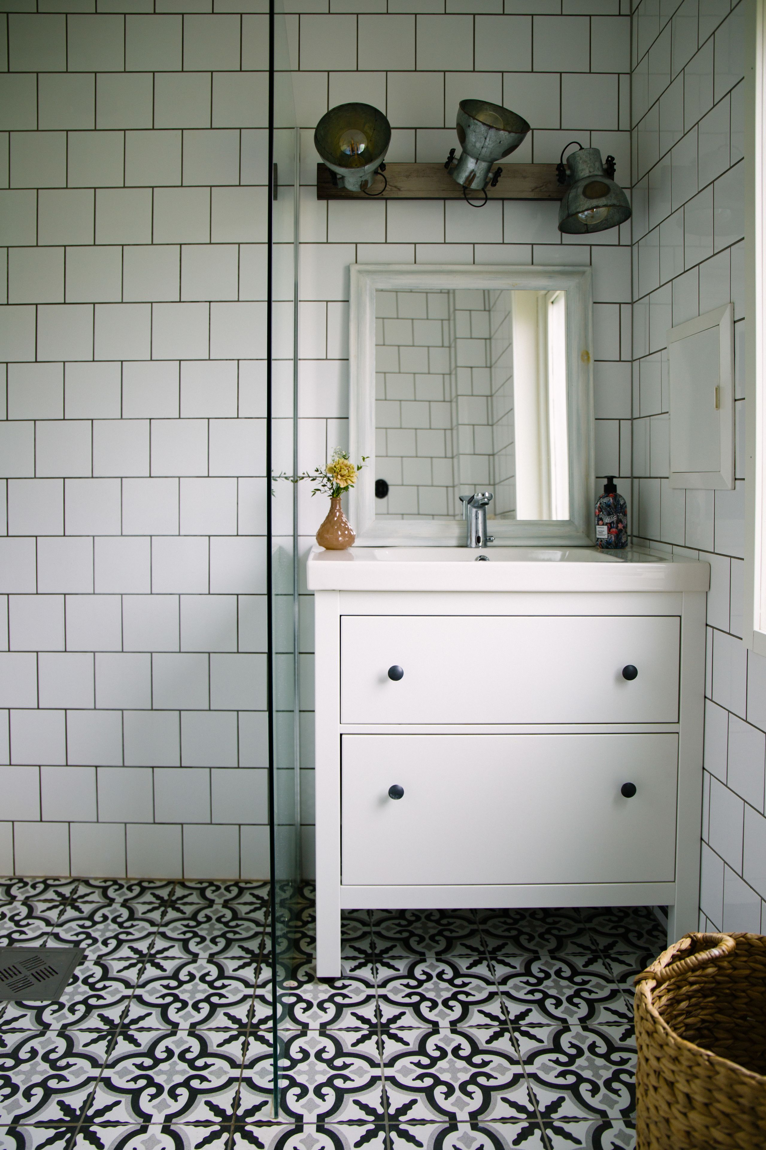 Un bagno con pareti piastrellate in bianco, un lavandino bianco con due cassetti e uno specchio sopra il lavandino. Sul lato sinistro del lavandino c'è un vaso con dei fiori.