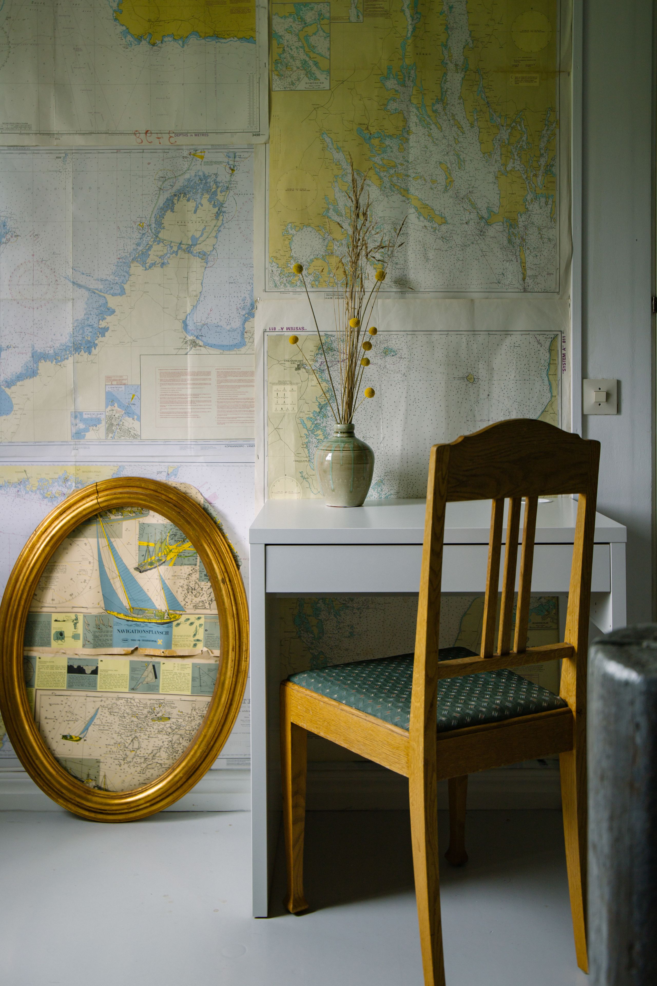 Una habitación con una silla de madera, un mapa en la pared y un jarrón con plantas secas.