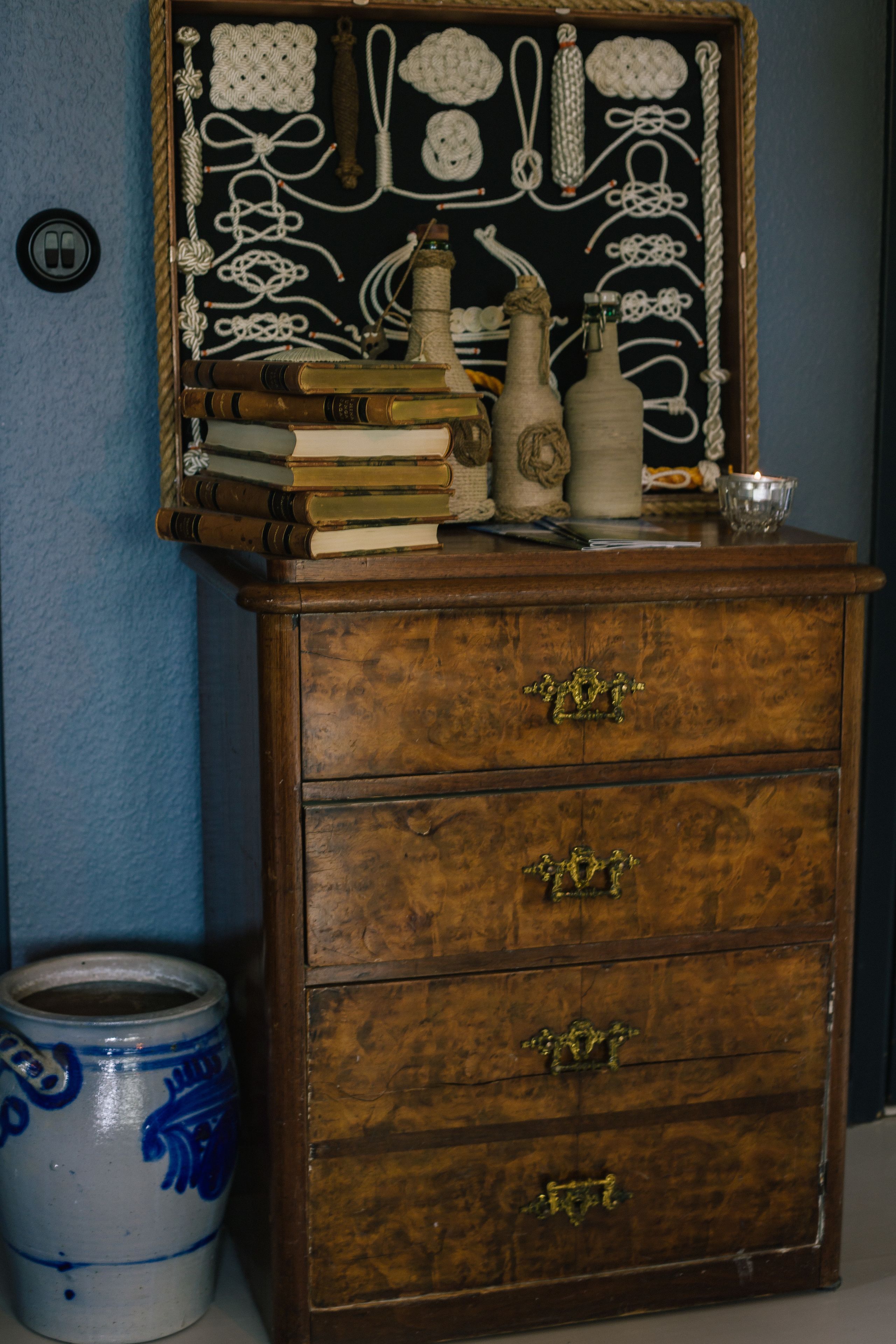 A wooden chest of drawers with brass handles, a blue vase, and a framed crochet pattern on the wall behind it.