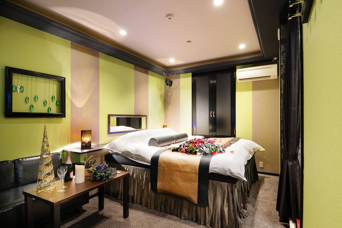 A hotel room with two beds, a small table with a bottle and glasses, a decorative tree, and a wall-mounted air conditioner.