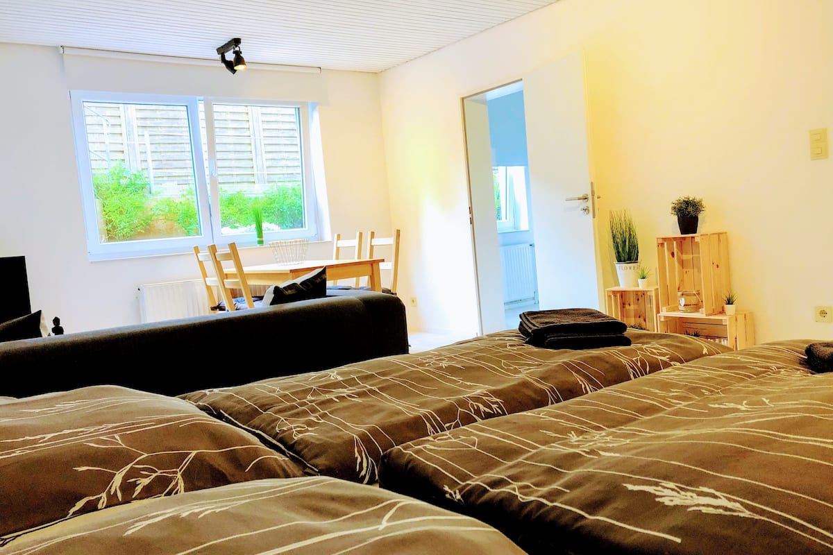 A bedroom with two beds, a window with blinds, a chair, a small table, and a door leading outside.