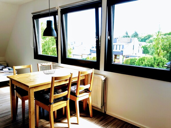 Un espace repas avec une table et des chaises en bois, un luminaire suspendu et une vue sur les bâtiments et la verdure, grâce à de grandes fenêtres.