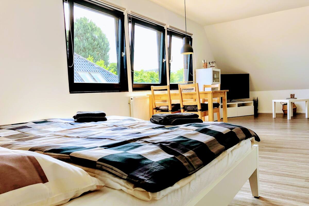 A bedroom with a bed, a desk, a chair, a TV, and large windows.
