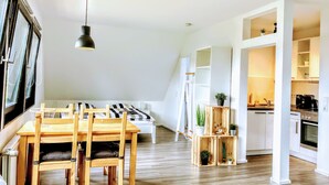 A modern kitchen with white cabinetry, a dining area with a wooden table and chairs, and a loft-style bedroom with a slanted ceiling. - Appartements Summer Break (Scharbeutz)