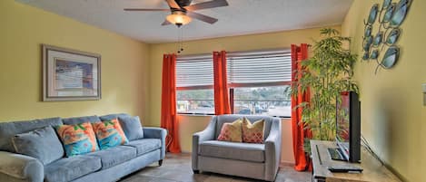 A living room with a grey sofa, a grey armchair, a ceiling fan, a framed picture on the wall, and a large window with red curtains.