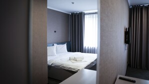 Una habitación de hotel con cama, televisor montado en la pared y ventana con cortinas.