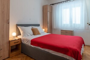 Une chambre à coucher comprenant un lit, des tables de chevet, une armoire et une fenêtre avec des rideaux légers.