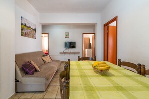 Un salon avec un canapé, une table à manger sur laquelle se trouve un saladier rempli de bananes, et un téléviseur fixé au mur.
