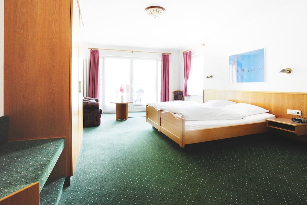 A hotel room with a bed, a desk, a chair, a wardrobe, and a view of the outside.