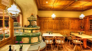 Un intérieur traditionnel en bois, avec un poêle central aux couleurs vert et or, des tables et des chaises en bois, et des luminaires suspendus.