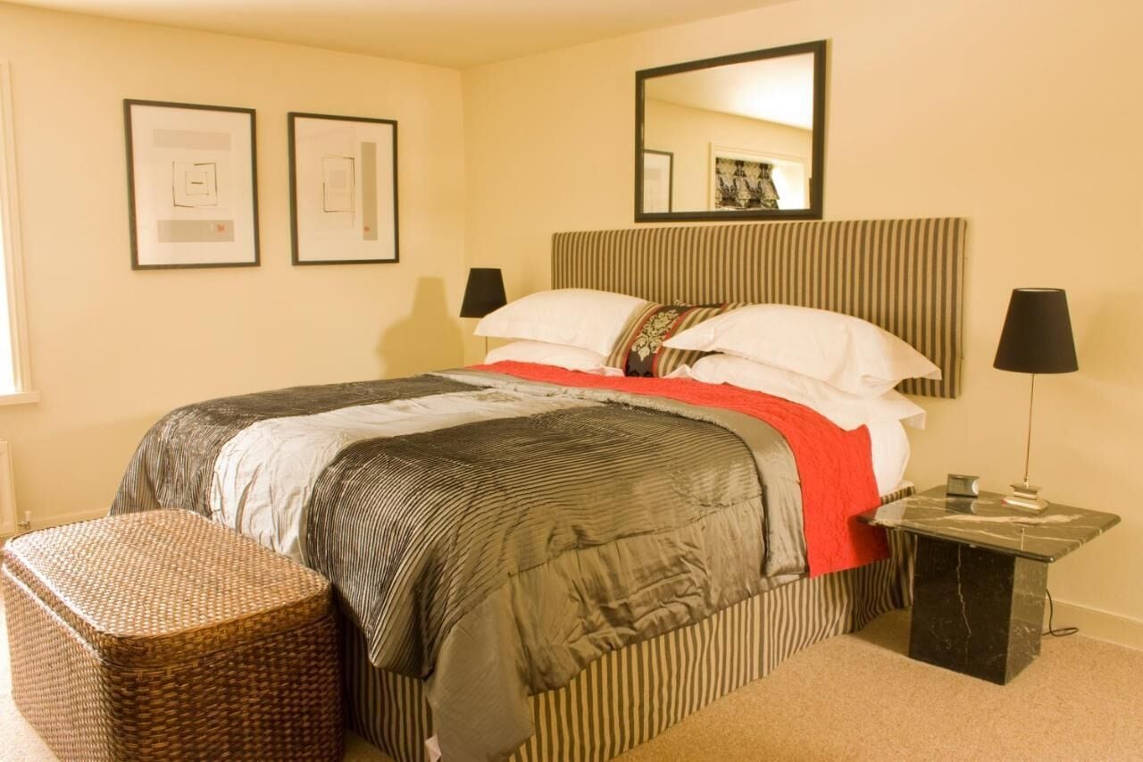 Un dormitorio ordenado con una cama, una mesita de noche, una lámpara y un taburete pequeño de mimbre.