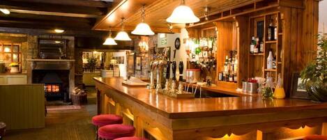 A bar area with wooden bar stools, a fireplace, and a variety of bottles and glasses on shelves.