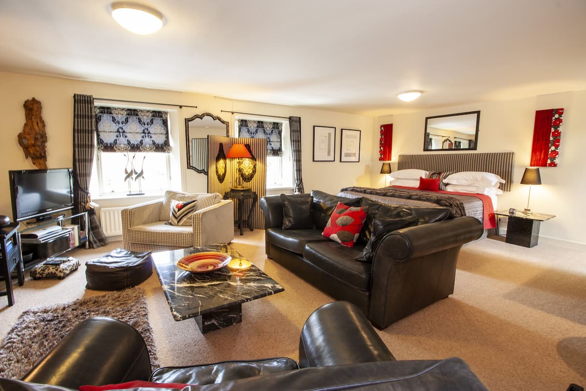 A modern living room with a sofa, armchair, and a television. There is a dining table with a lamp, and a bed with a red blanket in the background.