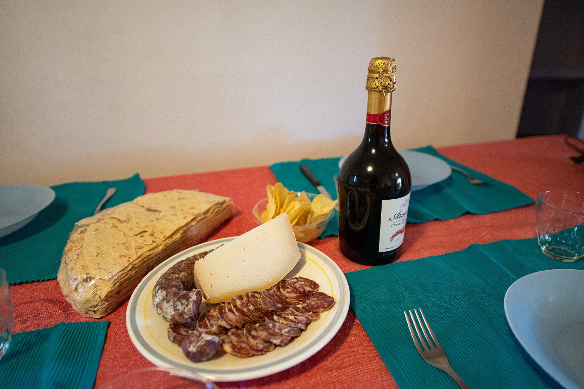Ein Tisch mit einer Weinflasche, einem Käse- und Wurstteller sowie einem verpackten Brot.