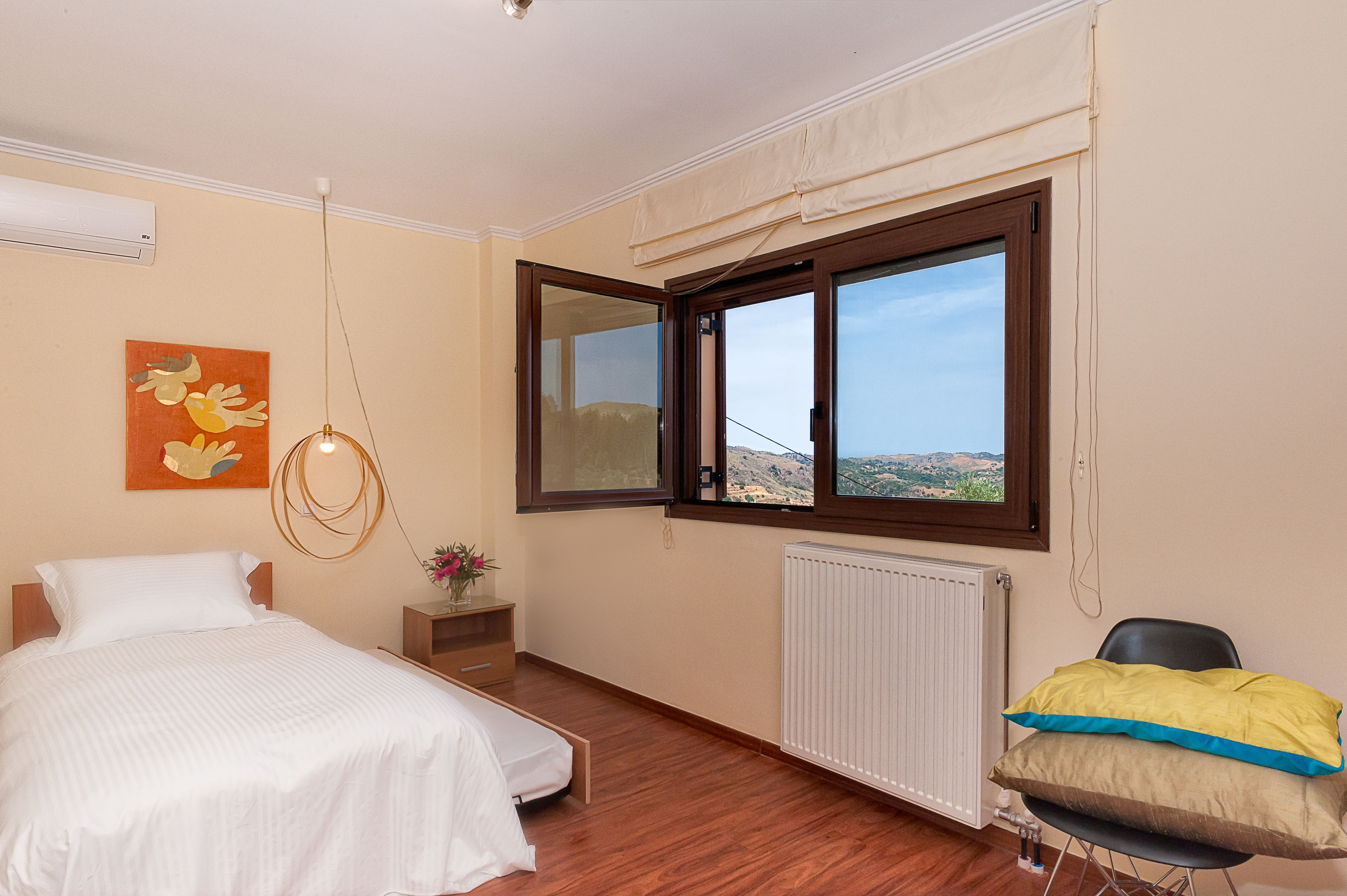 A bedroom with a bed, a chair, a window with a view, a radiator, and a wall-mounted air conditioner.