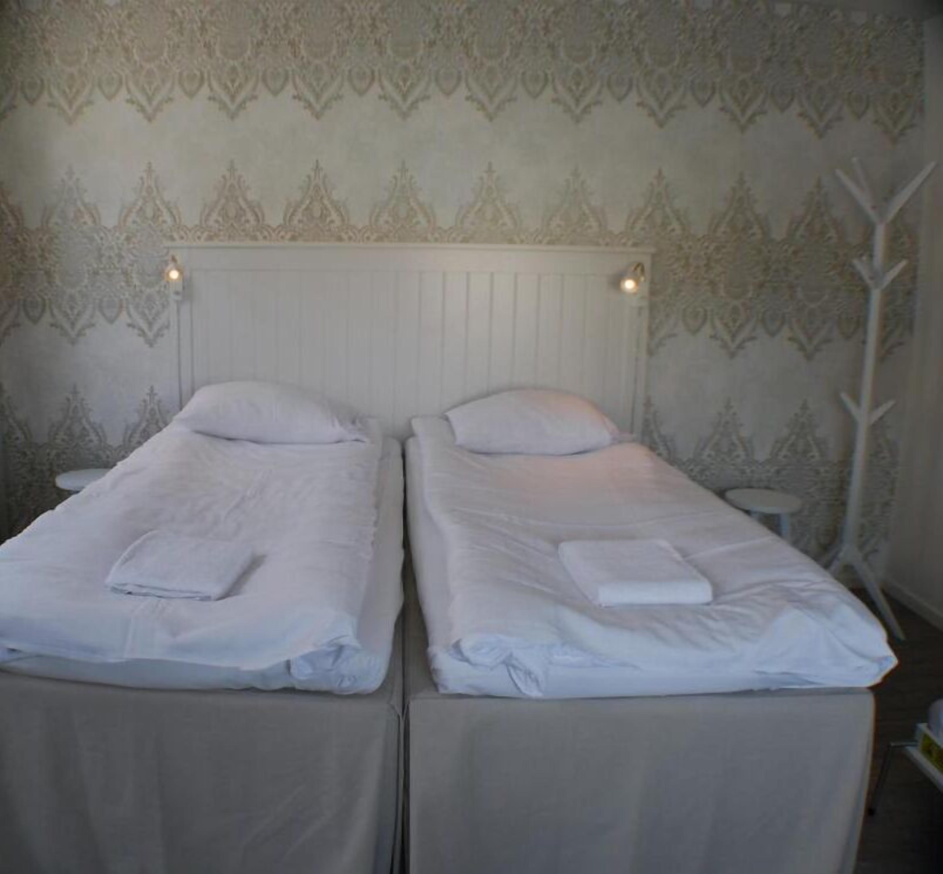 Una habitación con dos camas individuales, cada una con colcha y almohadas blancas, cabecero blanco y papel tapiz estampado.